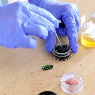 Person wearing blue gloves applying a substance from a small container onto a green object on a wooden surface.